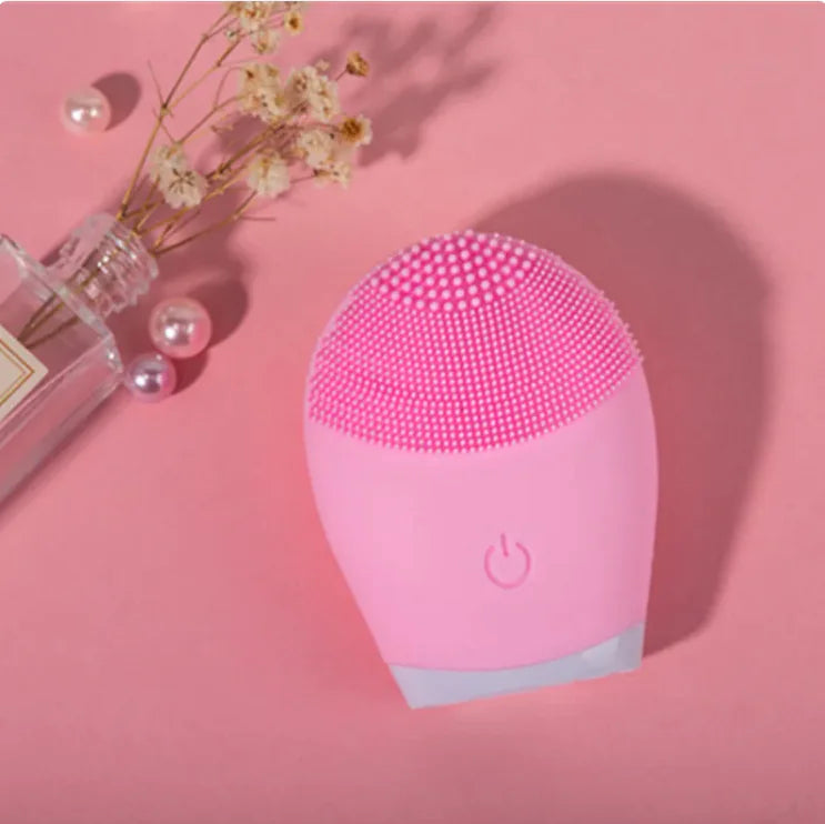 Pink facial cleansing brush on a pink background with a bottle and flowers.